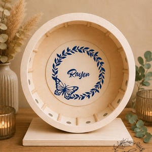 May include: A light-colored wooden wheel-shaped decorative item with a flat base. The center features a blue butterfly and the name "Rayen" within a blue wreath. The item is displayed on a wooden surface.