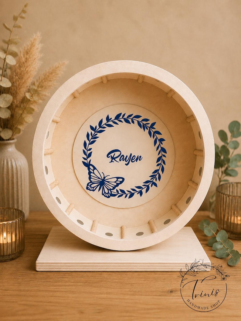 May include: A light-colored wooden wheel-shaped decorative item with a flat base. The center features a blue butterfly and the name "Rayen" within a blue wreath. The item is displayed on a wooden surface.