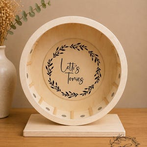 May include: A wooden hamster wheel with a light-colored finish. The wheel has a circular design with a flat base. The center of the wheel features the text "Lotti's Tones" inside a black leaf wreath. The wheel is designed for small pets.