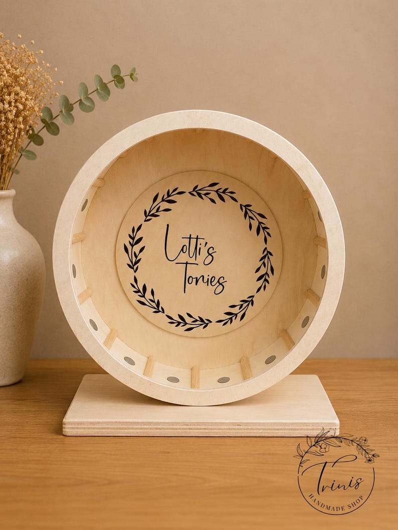 May include: A wooden hamster wheel with a light-colored finish. The wheel has a circular design with a flat base. The center of the wheel features the text "Lotti's Tones" inside a black leaf wreath. The wheel is designed for small pets.