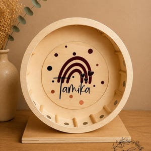 May include: A wooden hamster wheel with a personalized design. The wheel features a rainbow and heart design with the name "Tamika" in black script. The wheel is set on a wooden base and is a natural wood color.