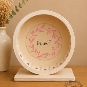 May include: A wooden hamster wheel with a light beige finish. The wheel features a pink floral wreath design with the name "Marie" in the center. The wheel is mounted on a rectangular wooden base. A vase with dried flowers is in the background.