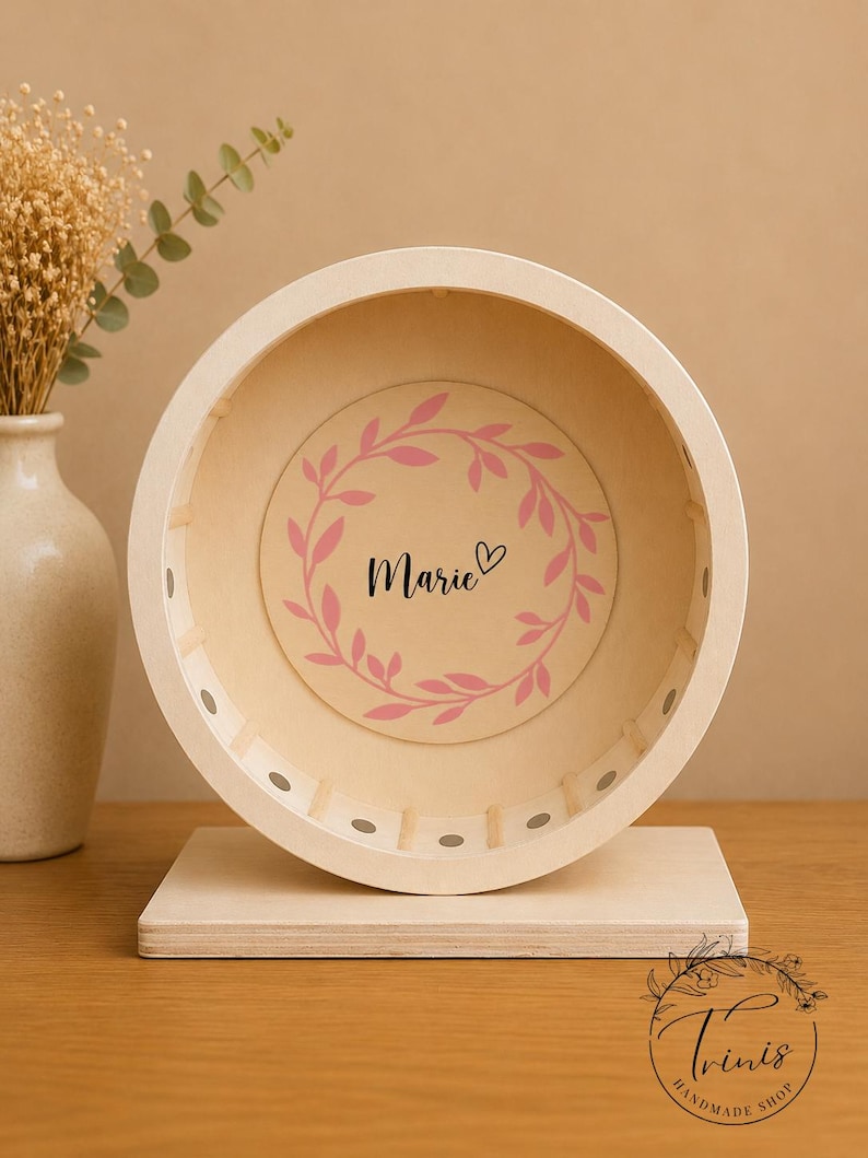 May include: A wooden hamster wheel with a light beige finish. The wheel features a pink floral wreath design with the name "Marie" in the center. The wheel is mounted on a rectangular wooden base. A vase with dried flowers is in the background.