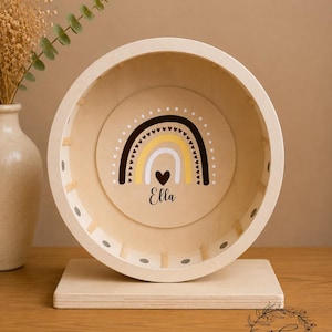 May include: A wooden hamster wheel with a rainbow design in the center. The rainbow features black, yellow, and white colors, with a heart and the name "Ella" below. The wheel is mounted on a wooden base.