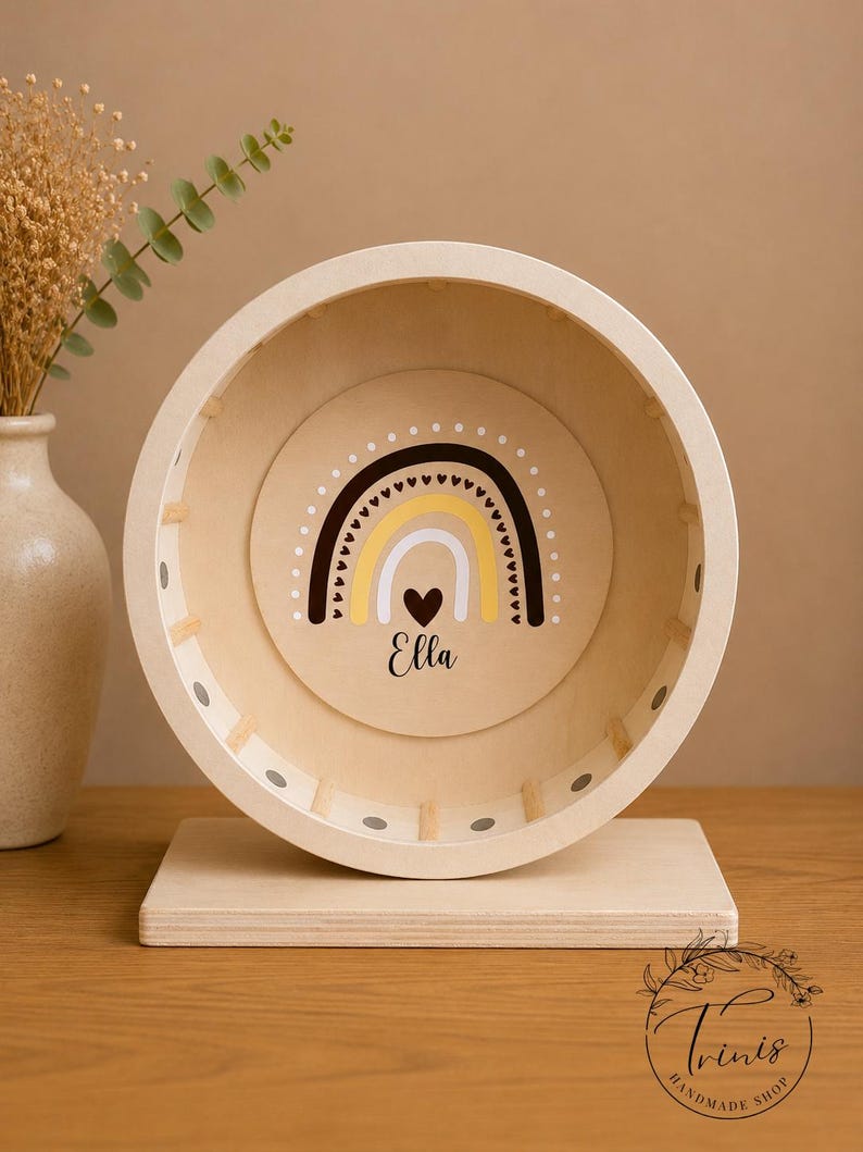 May include: A wooden hamster wheel with a rainbow design in the center. The rainbow features black, yellow, and white colors, with a heart and the name "Ella" below. The wheel is mounted on a wooden base.