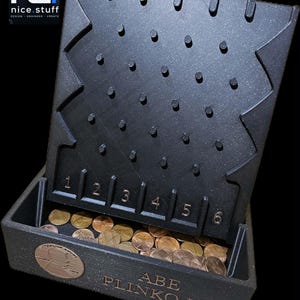 May include: A black Plinko game board featuring a grid of pegs, numbered slots one through six, and a collection of copper-colored pennies. The base displays the text ABE PLINKO.LN and a circular relief of a profile portrait of Abraham Lincoln.