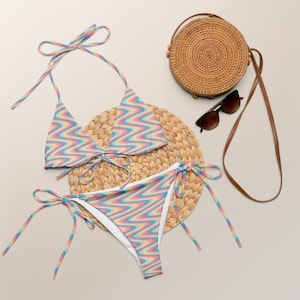 May include: A colorful swimsuit with a wavy pattern in pink, blue, and yellow. The swimsuit is on a woven straw mat with a round straw bag and sunglasses.