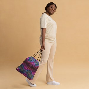 May include: A person wearing a light beige t-shirt and pants is holding a drawstring bag with a colorful geometric pattern. The bag has black drawstrings.