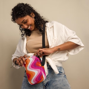 May include: A rectangular crossbody bag featuring a vibrant wavy pattern in shades of orange, pink, white, and light blue. The bag has a black adjustable strap and a black zipper closure, showcasing a bold, multi-coloured graphic design.