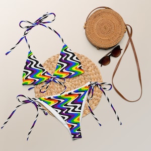 May include: A rainbow and black patterned bikini with a triangle top and a tie-side bottom. The bikini is on a woven straw mat with a round straw bag and sunglasses.