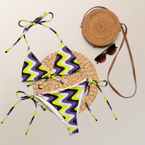 Puede incluir: Un bikini con estampado morado, amarillo y negro con un bolso de paja a juego y gafas de sol. La parte superior del bikini es de estilo triangular con tirantes ajustables y la parte inferior es de estilo atrevido con lazos en los lados.