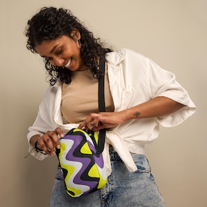May include: A rectangular crossbody bag featuring a vibrant wavy pattern in lime green, purple, black, and white. The bag has a black adjustable strap and a top zipper closure, constructed from a smooth material with a distinct graphic design.