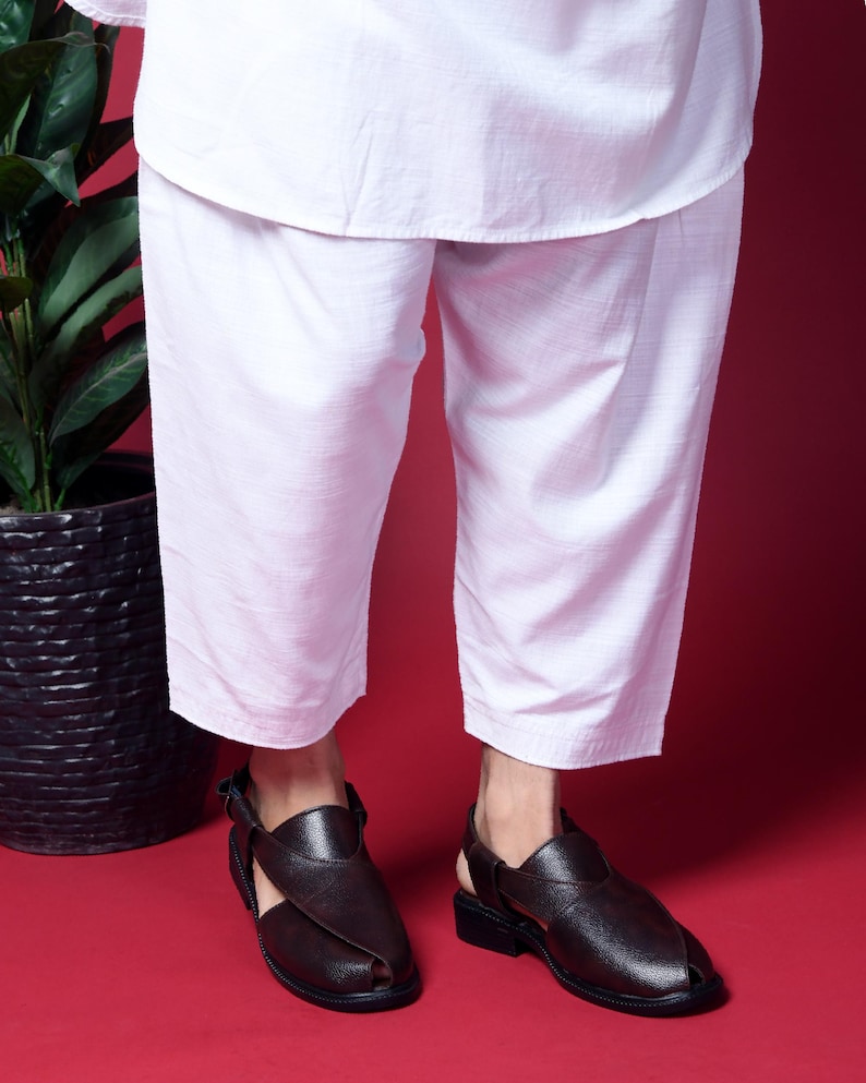 May include: A pair of brown leather sandals with a strap across the foot and an open toe. The sandals are worn by a person wearing white pants.