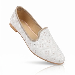 Può includere: Una scarpa piatta senza lacci di colore bianco con un motivo a rombi e dettagli floreali ricamati. La scarpa presenta un bordo beige lungo l'apertura e una suola piatta. Il design è composto da motivi quadrati ripetuti con stelle.