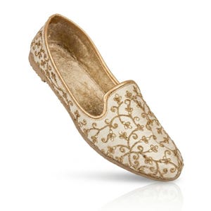 May include: Ivory-colored slip-on shoe with gold floral embroidery. The shoe features a rounded toe and a gold trim along the edges. The sole is a light brown color, and the interior appears to be a soft, golden fabric. A decorative footwear item.