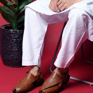 May include: A pair of brown leather sandals with a strap across the top of the foot and an open toe. The sandals have a black sole and are worn by a person sitting on a chair.