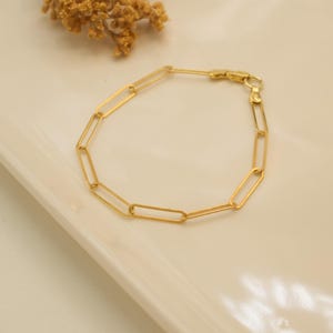 Könnte beinhalten: Ein goldenes Kettenarmband mit einem schlichten, rechteckigen Gliederdesign. Das Armband ist aus poliertem Metall gefertigt und hat einen sicheren Verschluss.