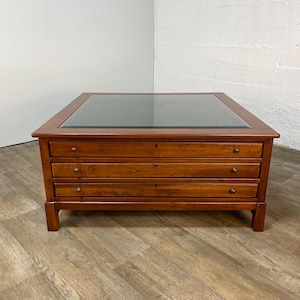 May include: A square wooden coffee table with a warm brown finish and a glass top insert. The table features three horizontal drawers stacked vertically, each equipped with two small, round metal knobs and a central keyhole escutcheon.