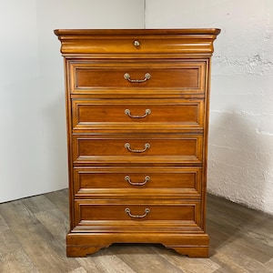May include: A wooden chest of drawers with five drawers and brass handles. The chest is a warm, medium brown color and has a decorative top and base. The drawers are framed with detailed molding. The chest is set against a white wall and a wooden floor.