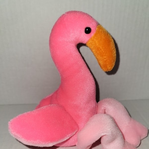 Może przedstawiać: Różowa pluszowa zabawka w kształcie flaminga z jasnopomarańczowym dziobem i czarnymi oczami. Flaming siedzi z podwiniętymi nogami.