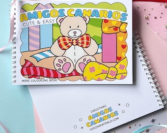 May include: A spiral-bound mini coloring book with the title "AMIGOS CANARIOS" and the words "CUTE & EASY." The cover features a cartoon teddy bear with a plaid bow tie. The background is a colorful illustration with gift boxes and bows.