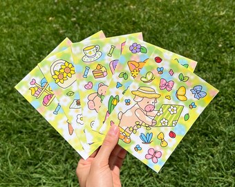Könnte beinhalten: Eine Hand hält einen Stapel bunter Aufkleber mit einem pastellfarbenen Blumen- und Tiermotiv. Die Aufkleber zeigen Illustrationen von Sonnenblumen, Teetassen, Teddybären, Schmetterlingen und einem Schwein mit Hut. Der Hintergrund ist ein weiches Grün.