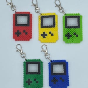 以下が含まれることがあります： 携帯用ビデオゲーム機の形をした、カラフルなピクセルアートのキーホルダーチャームが5つ。チャームは赤、黄、緑、青です。各チャームには、白黒のピクセルデザインが施されています。