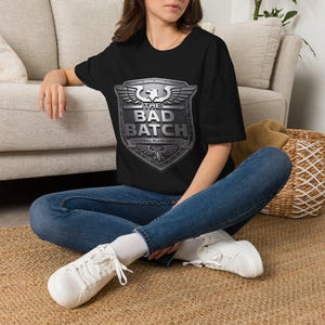 May include: A black short-sleeved t-shirt featuring a large, metallic-toned shield graphic on the chest. The shield displays a winged eagle emblem at the top and bottom, with the text THE BAD BATCH and FEDERAL ELITE UNIT printed in the center.