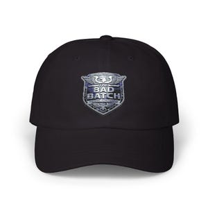 May include: A black baseball cap featuring a centered, metallic-toned shield emblem. The emblem displays a winged crest at the top and the text THE BAD BATCH in bold, silver-colored lettering, set against a dark, textured, and weathered background.