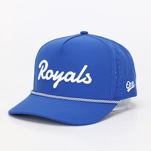May include: A blue baseball cap with the word "Royals" in white script across the front. The cap has a curved brim, a white and blue rope detail, and a small logo on the side. The cap is a sports accessory.