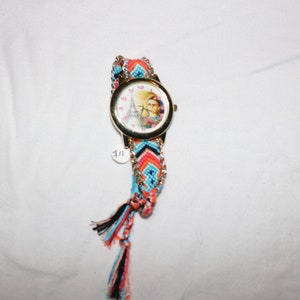 Op de afbeelding: Een kleurrijk geweven armbandhorloge met een gouden kast en een witte wijzerplaat met een afbeelding van de Eiffeltoren. Het horloge heeft een roze, blauwe en oranje geweven band met zwarte kwastjes.