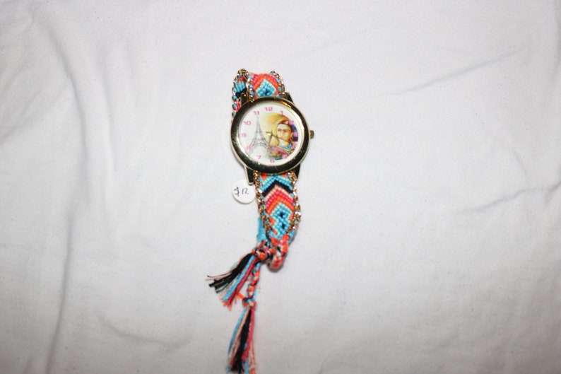 Op de afbeelding: Een kleurrijk geweven armbandhorloge met een gouden kast en een witte wijzerplaat met een afbeelding van de Eiffeltoren. Het horloge heeft een roze, blauwe en oranje geweven band met zwarte kwastjes.