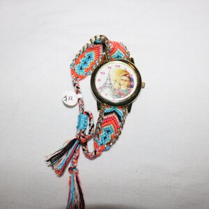 Op de afbeelding: Een gouden polshorloge met een witte wijzerplaat met een kleurrijk beeld van de Eiffeltoren en een vrouw. Het horloge is bevestigd aan een kleurrijk geweven armband met kwastjes.
