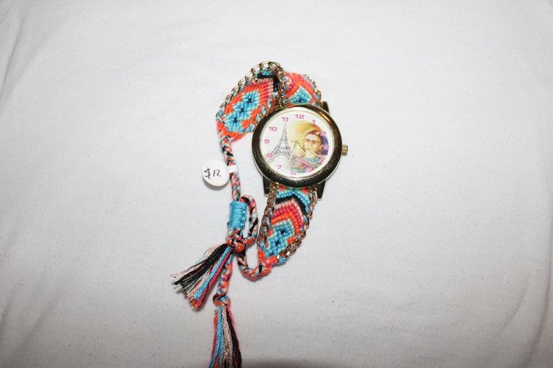 Op de afbeelding: Een gouden polshorloge met een witte wijzerplaat met een kleurrijk beeld van de Eiffeltoren en een vrouw. Het horloge is bevestigd aan een kleurrijk geweven armband met kwastjes.