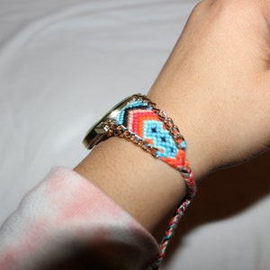 Op de afbeelding: Een gouden horloge met een kleurrijk geweven armband. De armband is gemaakt van blauwe, oranje en rode draad en heeft een gouden ketting die erin is geweven.