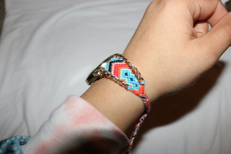 Op de afbeelding: Een gouden horloge met een kleurrijk geweven armband. De armband is gemaakt van blauwe, oranje en rode draad en heeft een gouden ketting die erin is geweven.