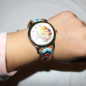 Op de afbeelding: Een gouden polshorloge met een witte wijzerplaat met een kleurrijk beeld van de Eiffeltoren en een vrouw. Het horloge heeft een veelkleurige geweven band.