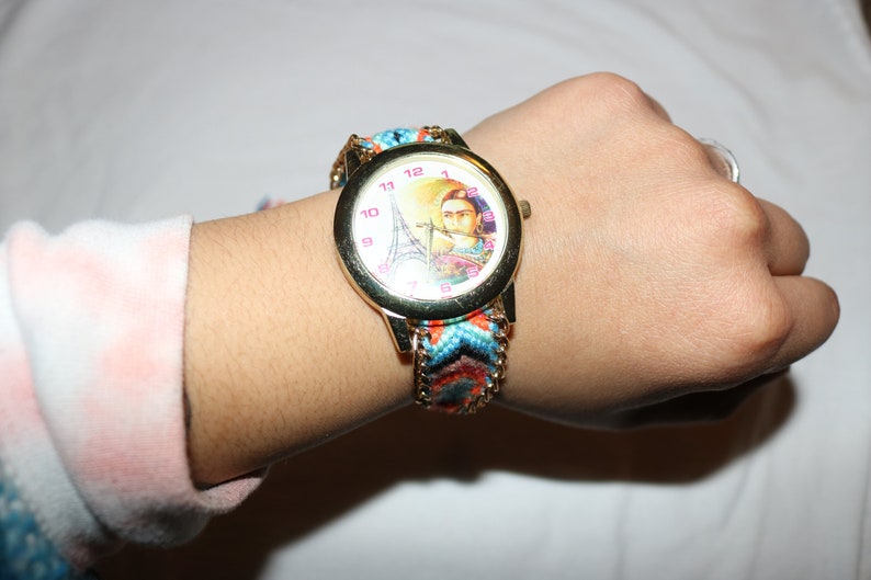 Op de afbeelding: Een gouden polshorloge met een witte wijzerplaat met een kleurrijk beeld van de Eiffeltoren en een vrouw. Het horloge heeft een veelkleurige geweven band.