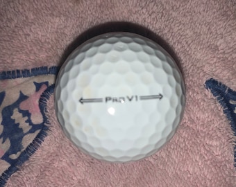 May include: A white golf ball with the text "Pro V1" printed on it. The ball has dimples and two arrows pointing in opposite directions.