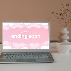 Op de afbeelding: Een laptop met een roze en wit scherm waarop de tekst "stream is ending soon" staat met een wolkenachtergrond. De laptop staat op een witte tafel met een vaas met gedroogde bloemen en een witte kom.