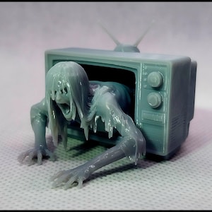 May include: A light blue figurine of a vintage television set with a ghostly figure emerging from the screen. The figure has long hair and is in a crawling position. The television has two knobs and an antenna.