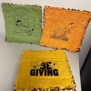 May include: Three square, decorative fabric pieces with frayed edges. One is green with a Snoopy and Woodstock graphic and text. Another is orange with a Snoopy graphic. The third is yellow with Snoopy, Woodstock, and the words "BE GIVING".