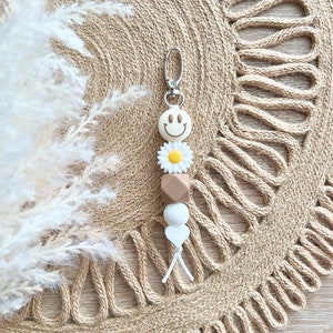 May include: A white and brown silicone keychain with a smiley face, a white daisy, a brown bead, a white bead, and a white heart charm. The keychain is attached to a silver key ring.
