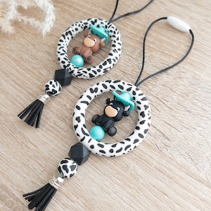 May include: Two black and white cow print silicone teethers with a black and brown bull and a black bull wearing a blue hat. Each teether has a black tassel and a turquoise bead.