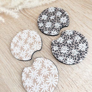 May include: Three round car coasters with a beige background and white floral designs. Two coasters feature a leopard print pattern with white flowers. One coaster features a smaller white flower pattern.