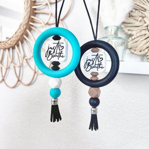 May include: Two car air fresheners. One is teal, the other is navy blue. Each has a circular ring, a square with the words "just breathe", and a tassel. The teal one has white, black, and teal beads. The navy one has brown and navy beads.