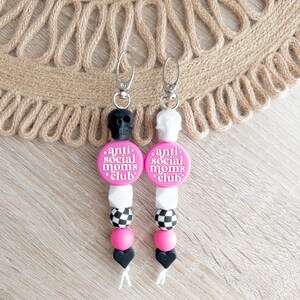 Puede incluir: Dos llaveros de silicona rosa y negro con un dije de calavera en la parte superior. Los dijes tienen cuentas de cuadros negros y blancos, cuentas rosas y una cuenta de coraz&oacute;n negra. Los dijes tienen un c&iacute;rculo rosa con el texto "anti social moms club" en &eacute;l.