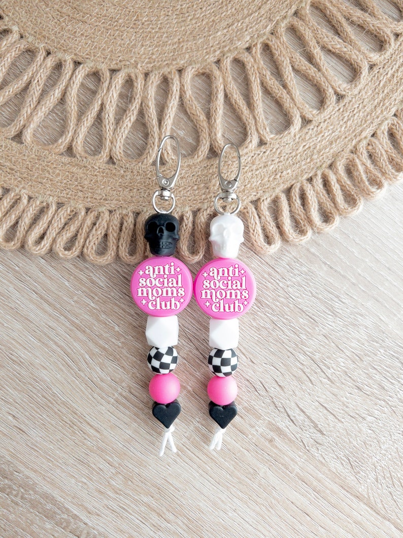 Puede incluir: Dos llaveros de silicona rosa y negro con un dije de calavera en la parte superior. Los dijes tienen cuentas de cuadros negros y blancos, cuentas rosas y una cuenta de coraz&oacute;n negra. Los dijes tienen un c&iacute;rculo rosa con el texto "anti social moms club" en &eacute;l.