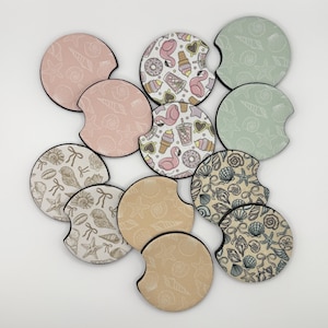 May include: A collection of car coasters with various designs. Some coasters feature seashell patterns in pink, beige, and green. Others display flamingo, ice cream, and donut motifs. Each coaster has a black border.