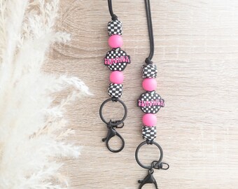 Puede incluir: Dos cordones negros y blancos a cuadros con cuentas rosadas y un dije de corazón rosa con la palabra "Teacher" en él. Cada cordón tiene un clip de metal negro.